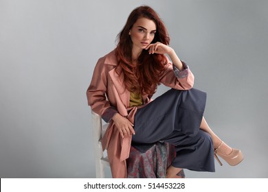 Fashion Model Style. Fashionable Woman In Stylish Clothes Posing On Grey Background In Studio. Beautiful Sexy Girl Wearing Pink Coat, Purple Trousers, Beige Shoes Sitting On Chair. High Resolution