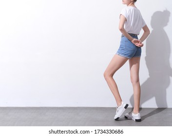 Fashion model in t-white shirt with legs wear in blue jeans shorts  and white sneakerstanding on gray background with shadow

