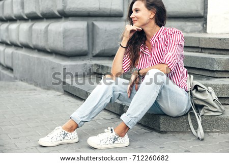 Fashion model wearing ripped boyfriend jeans, red striped shirt, sneakers and backpack posing in the city street. Fashion urban outfit. Casual everyday clothing style.