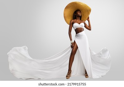 Fashion Model in White Dress and Big Summer Hat. Sexy Stylish Woman in Long Slit Gown flying on Wind showing tanned Leg over isolated Studio Background