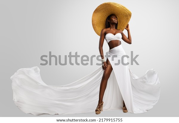 Fashion Model in White Dress and Big Summer\
Hat. Sexy Stylish Woman in Long Slit Gown flying on Wind showing\
tanned Leg over isolated Studio\
Background