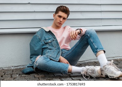 Fashion Model Young Man In Stylish Denim Jacket In Ripped Fashionable Blue Jeans In Trendy White Sneakers Sit On Stone Tile Near Wall In City. Handsome Guy In Vintage Casual Clothes On Street Outdoors