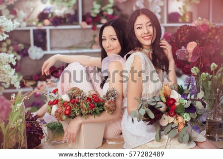 Fashion models in tender dresses posing and looking at camera. Beautiful asian florist girls making bouquet of flowers on table for sale against floral bokeh background in flower shop indoors. Two
