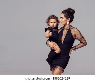 Fashion Mom In Black Elegant Dress And Oriental Caucasian Kid. Studio Gray Background Photo Shoot Style Exquisitely Festive Feminism. Holiday New Year Mother's Day Outfit  Daughter Girl Boy Son Baby