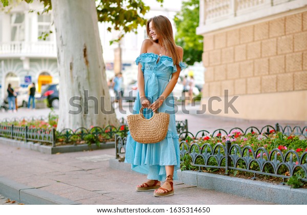Fashion outdoor\
full length image of stylish elegant woman wearing pretty trendy\
blue maxi dress and gladiator sandals, holding straw bag, summer\
spring time, tourist in\
Europe.