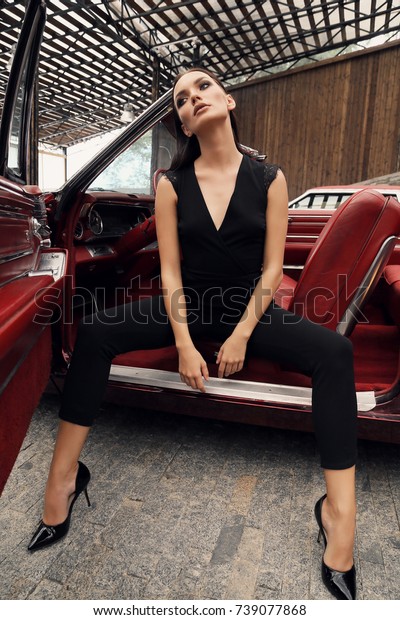 fashion outdoor photo\
of beautiful girl with dark hair in elegant clothes posing in\
luxurious red retro car