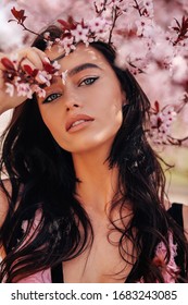 fashion outdoor photo of beautiful woman with dark hair in elegant dress with accessories posing among blossoming peach trees