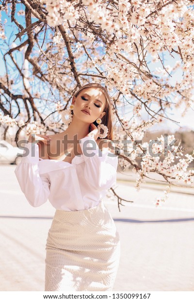 美しい金髪の美しい若い女性と優雅なドレスを着た美しい若い女性のアウトドア写真 花をつける桃の木を持つ庭にポーズをとる豪華なイヤリング の写真素材 今すぐ編集