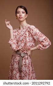 Fashion Photo Of A Beautiful Elegant Young Asian Woman In A Pretty Beige Dress With A Floral Pattern Posing Over Brown Coffee Background. Studio Shot. Portrait