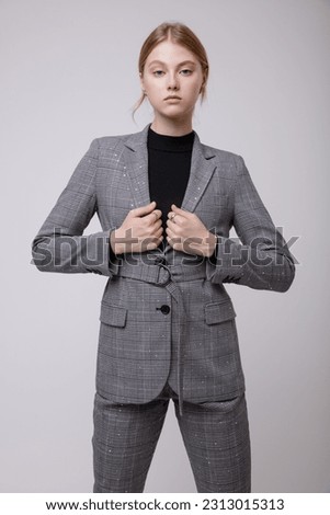 Fashion photo of a beautiful elegant young woman in a pretty gray suit, pants, trousers, jacket, blazer, black top, pearl choker posing over white, soft gray background. Studio Shot, portrait. Blonde