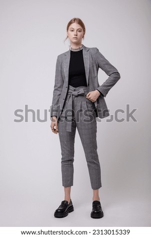 Fashion photo of a beautiful elegant young woman in a pretty gray suit, pants, trousers, jacket, blazer, black top, pearl choker posing over white, soft gray background. Studio Shot, portrait. Blonde