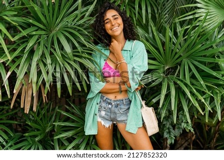 Fashion photo of pretty woman in stylish summer outfit posing over tropical  garden. Wearing  straw bag and   bracelets.