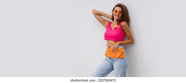 Fashion Photo Of Sensual  Woman In Colorful Summer Outfit , Pink Crop Top And Blue Jeans  Posing Over White Background. Holding Orange Bag. Summer Holidays