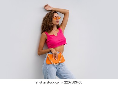 Fashion Photo Of Sensual  Woman In Colorful Summer Outfit , Pink Crop Top And Blue Jeans  Posing Over White Background. Holding Orange Bag. Summer Holidays