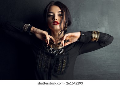 Fashion photo of sexy Indian woman with dark hair and bright make-up wearing black shirt and  luxurious gold necklace,bracelets, earrings posing at studio