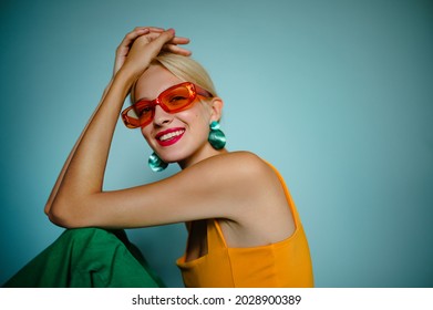 Fashion portrait of beautiful happy smiling model, woman wearing trendy orange sunglasses, green shell earrings, posing on blue background. Copy, empty space for text