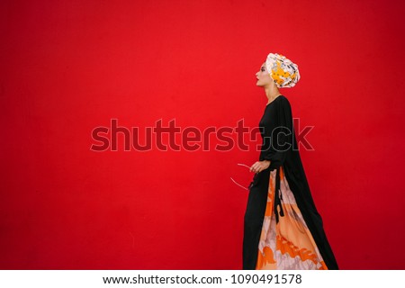 Fashion portrait of a beautiful, tall and fashionable woman of Middle Eastern descent. She is posing against a red, plain background. 