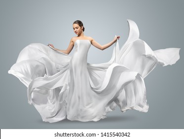Fashion portrait of a beautiful woman in a waving white dress. Light fabric flies in the wind. Light gray background. Girl posing in studio