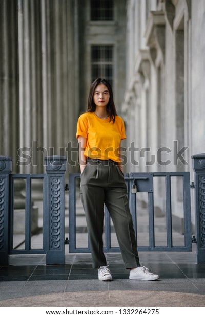 美しく 若く 優雅な中国のアジアの女の子のスマートなカジュアルな服装のファッションポートレート 彼女は日中 灰色の柱に黄色のティー オリーブグリーンのズボン 白いスニーカーをはいている の写真素材 今すぐ編集