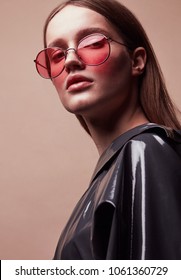 Fashion portrait of beautiful young woman in round red sunglasses and grey latex jacket posing in studio