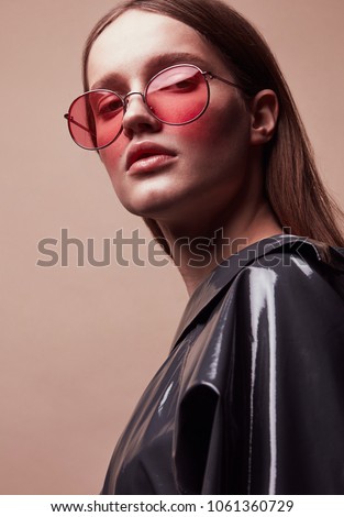 Fashion portrait of beautiful young woman in round red sunglasses and grey latex jacket posing in studio
