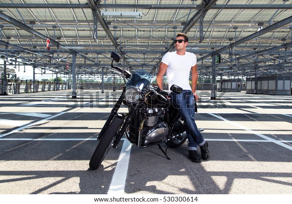 Fashion portrait of biker man wearing a white\
shirt, blue jeans and sunglasses sitting on his classic motorcycle\
in a empty car parking