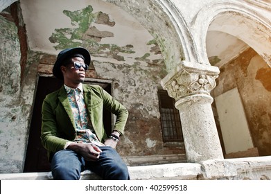 Fashion Portrait Of Black African American Man On Green Velvet Jacket And Black Hat, Sitting On The Railing Background Old Vintage House With Arch 
