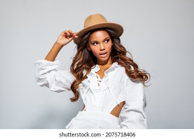 Fashion Portrait Black Woman in white stylish linen dress. Makeup, curly Hair. Luxury Fashion model African American posing in studio against a light wall. Beautiful Black Woman in beige felt Hat     
