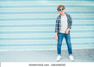 Fashion Portrait Of Caucasian Blonde Hair 12 Year Old Teenager Boy Dressed T-shirt And Checkered Shirt In Blue Sunglasses Posing On Turquoise Blue Background Wall Background. Teens Fashion Concept.