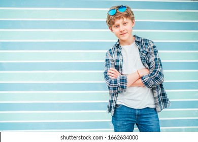 Fashion Portrait Of Caucasian Blue-eyed Blonde Hair 12 Year Old Teenager Boy Dressed T-shirt And Checkered Shirt With Sunglasses Posing On Turquoise Blue Background Wall Background. Teens Fashion.