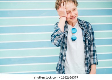 Fashion Portrait Of Caucasian Blue-eyed Blonde Hair 12 Year Old Teenager Boy Dressed T-shirt And Checkered Shirt With Sunglasses Cramped Because Of Shooting Turquoise Blue Background Wall Background