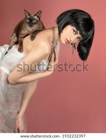 Fashion portrait of a dark-haired woman with short hair with a cat on her back on a pink background