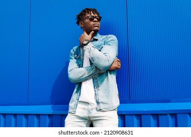 Fashion portrait of a handsome smiling stylish dark-skinned man, dressed in a jacket and jeans. Fashionable man posing against blue wall in sunglasses - Powered by Shutterstock