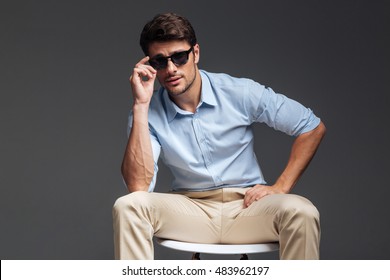 Fashion Portrait Of Handsome Young Man In Sunglasses Sitting On The Chair Over Grey Background