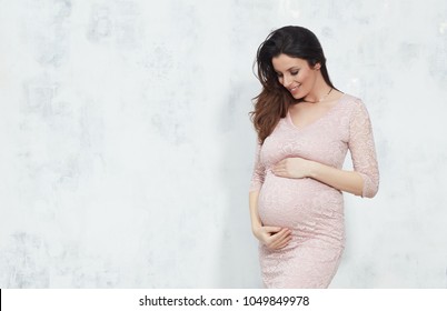 Fashion portrait of happy pregnant woman in elegant dress against white wall with copyspace.