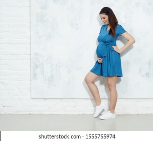 Fashion portrait of happy pregnant woman in blue summer dress against white wall with copyspace.