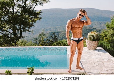 Fashion Portrait Of A Muscular Man In Swim Trunks At Swimming Pool. Rich Muscular Handsome Man Relax At Swimming Pool Outdoors. Luxury Lifestyle