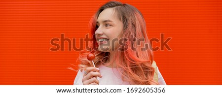 Fashion portrait of pretty smiling hipster woman with red flying haies and lollipop against the colorful orange wall