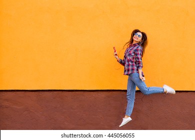 Fashion Portrait Of Pretty Smiling And Jumping Woman In Sunglasses With Smartphone Against The Colorful Orange Wall. Copyspace