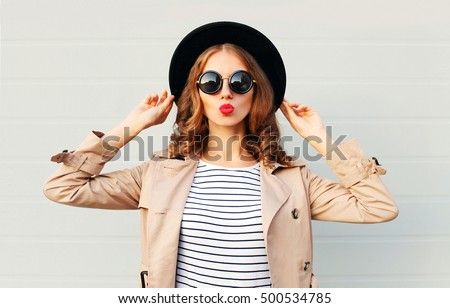 Fashion portrait pretty sweet young woman blowing red lips wearing a black hat sunglasses coat over grey background