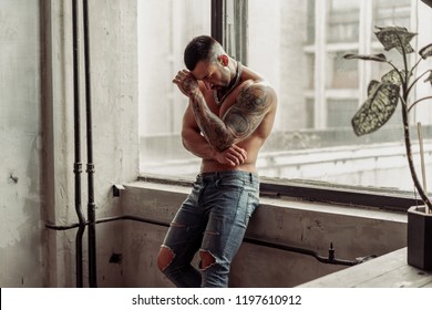 Fashion portrait of Sexy naked male model with tattoo and a black beard standing in hot pose on near the window. Loft room interior with grey concrete wall. Professional Studio image.