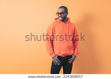 Fashion portrait of smiling handsome dark-skinned elegant man wearing hoodie with hat and jeans. Fashionable man posing near an orange wall in sunglasses