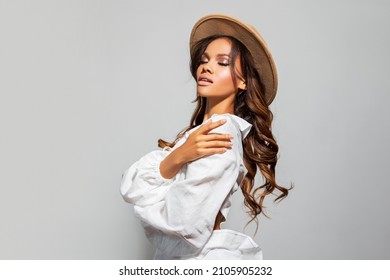 Fashion Portrait of stylish black woman in a Hat. Makeup-beige Lips. Beautiful model African American girl posing in a white shirt and beige stylish hat , long curly hair flowing on a gray background 