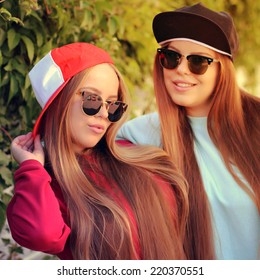 Fashion portrait of two pretty girls, wearing sweatshirt swag hats and sunglasses, having long blonde hairs outdoors on sunny summer day. Modern lifestyle concept. Photo toned style Instagram filters.
