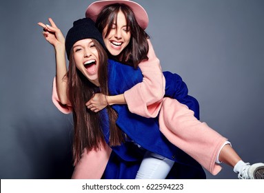 Fashion Portrait Of Two Smiling Brunette Women Models In Summer Casual Hipster Overcoat Posing On Gray Background. Girls Holding Each Other On Back 