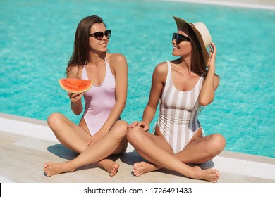 Fashion portrait of two stunning girls laying near pool, amazing long blonde and brunette hair, stylish bikinis, pretty faces tanned slim body,eating watermelon , vacation