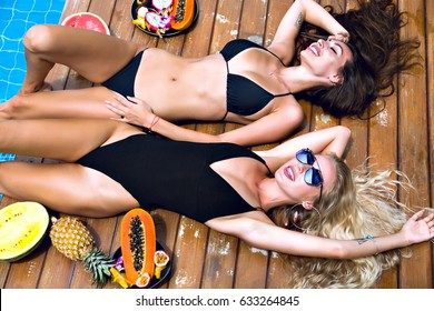 Fashion portrait of two stunning sexy girls laying near pool, amazing long blonde and brunette hair, stylish black bikinis, pretty faces tanned slim body, a lot of tropical exotic fruits, vacation.