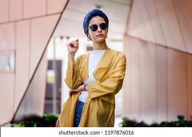 A fashion portrait of a young and attractive Muslim Malay woman in the city. She is well-dressed and fashionable with a navy turban (hijab head scarf) and sunglasses; she is posing for her photograph.