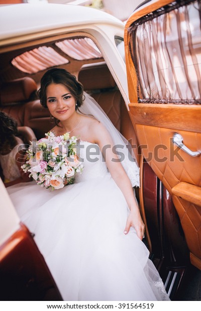 Fashion portrait of young bride with white\
dress with bouquet near retro car in\
spring