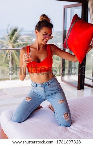 Fashion portrait of young fit woman with abs in blue jeans and red top, big breast, starts morning in her bedroom at villa with amazing tropical view. Holds glass of water.